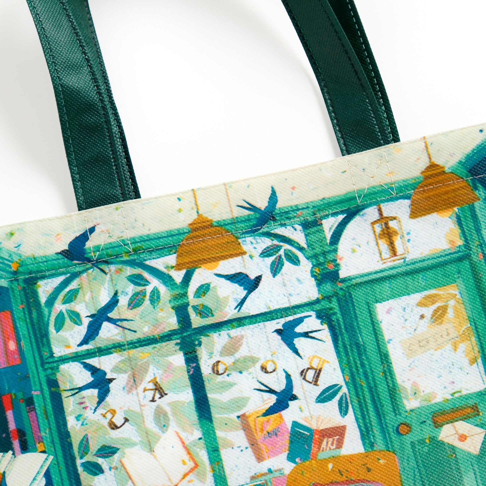 The Bookshop Afternoon Reusable Shopping Bag by Victoria Ball features a vibrant, illustrated scene with blue birds, hanging lights, leafy patterns, and windows. This sustainable tote has green handles and a whimsical, colorful design.
